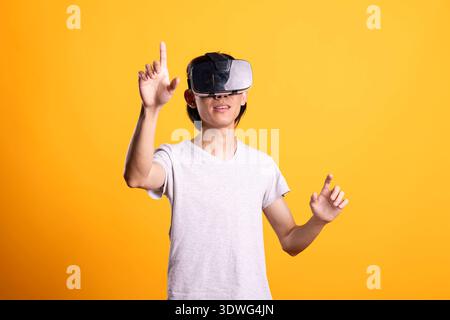 Curious young asian man wears VR headset, participating in interactive gaming simulation. Focused male individual immersed in virtual reality world, selecting digital elements with index finger. Stock Photo