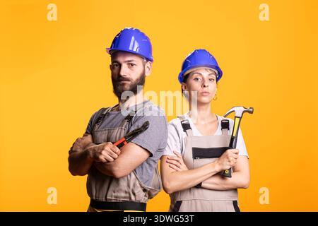 Caucasian builders pose in overalls with arms crossed expressing readiness for repair tasks. Team of renovators with tools in hand, facing camera in studio, suitable for handyman service advertising. Stock Photo
