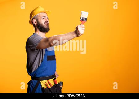 Male contractor in hardhat extends paintbrush forward, appearing to inspect coverage or gesture for paint direction. White repairman stands representing painting work or home renovation services. Stock Photo