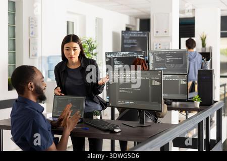 Adult programmers review website functionality together, comparing code on computer screen in startup office. Focused discussion reflects teamwork, innovation and software development culture. Stock Photo