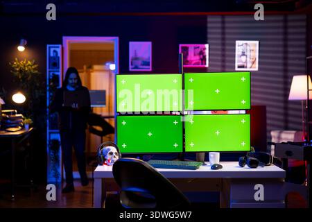 Hacker entering RGB lit apartment with mockup multi monitor setup, working on building stealth malware. Cybercriminal coming home to underground HQ, using laptop to get past firewalls Stock Photo