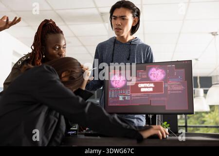Professional programmers gather around monitors, reacting to sudden system error alert. Intense office moment shows mixed emotions as diverse team confronts frustrating software malfunction. Stock Photo