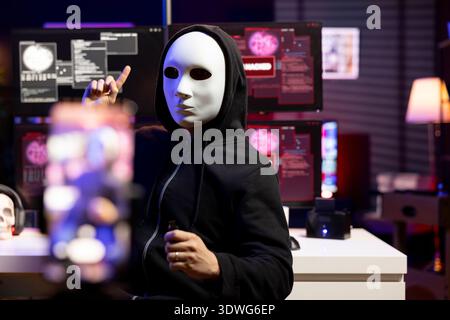 Hacker hidden in neon lit hideout films video ultimatum with smartphone, sending it to foreign governments. Masked evil man threatens retaliation against foreign military targets Stock Photo