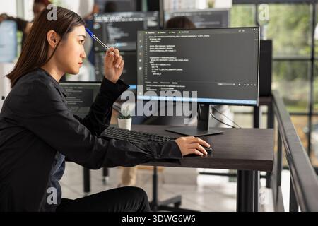 Asian female software engineer taps pen thoughtfully while analyzing code on desktop monitor, focused on web development and digital innovation. Adult woman programmer working on dynamic application. Stock Photo