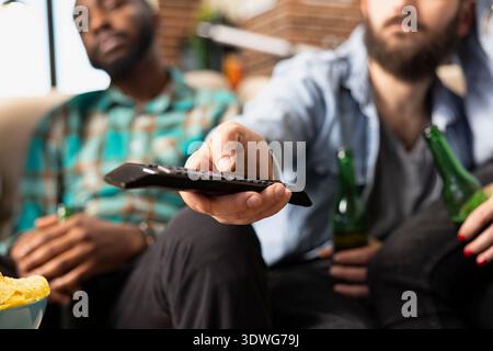 White bearded man operating television remote, changing volume and channels, with friends sitting on couch close by. Male individual uses device in picking show to watch during casual hangout at home. Stock Photo