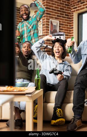 Cheerful multiethnic friends play online video games together, celebrating a win. Caucasian woman wears VR headset as gaming buddies watch and react, enjoying fun moment at home. Stock Photo