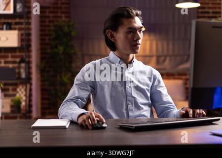 Asian man works remotely during evening hours, researching strategies and improving plans for small business. Freelancer concentrates on pc screen, typing and reviewing marketing data in home office. Stock Photo
