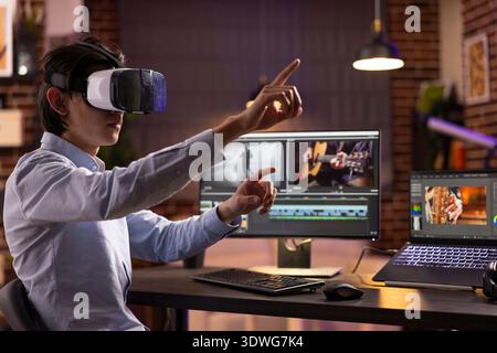 Self employed man editing immersive film montage using laptop and desktop pc. Asian male freelancer wearing VR glasses, explores digital visual effects and storytelling in creative home workspace. Stock Photo