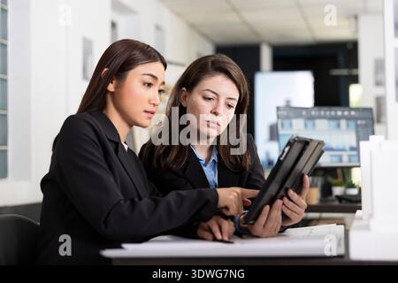 Caucasian and Asian women review architectural floor plans and documents on tablet. Female professionals work together on accurate building measurements and preparing construction project reports. Stock Photo