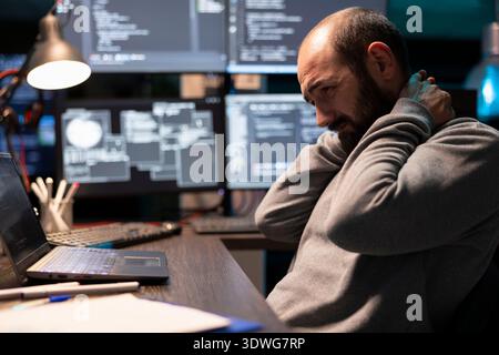 Stressed male programmer rubbing his neck after long hours of coding in front of multiple glowing monitors. The posture reflects workspace fatigue and physical strain of tech work. Stock Photo