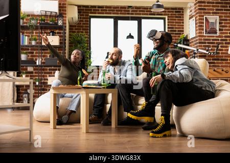 Cheerful group of friends enjoys weekend VR gaming in modern apartment. Diverse people engage in virtual reality with drinks and snacks, bonding in cozy living room during fun leisure time. Stock Photo