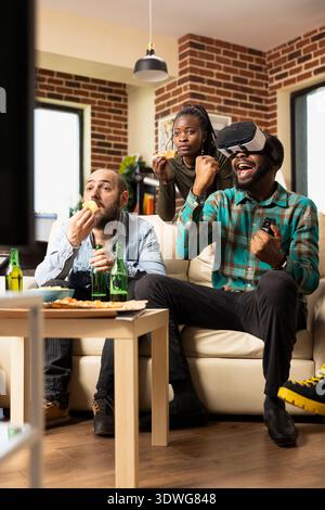 Black man wearing vr glasses celebrates gaming victory as friends eat snacks and pizza on couch. Diverse group enjoys weekend, cheering for african american male winning virtual reality tournament. Stock Photo