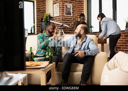 Cheerful male friends celebrate thrilling video game moment, giving high five while sitting on living room couch. Excited multiethnic men enjoying entertainment in modern apartment. Stock Photo