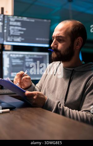 Programmer reviews paperwork with pen cap held between lips, checking coding scripts on laptop. Developer analyzes technical details on clipboard and confirms information against data on device. Stock Photo