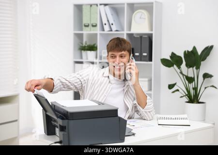 Smiling man working with printer and talking by smartphone at white desk in office Stock Photo