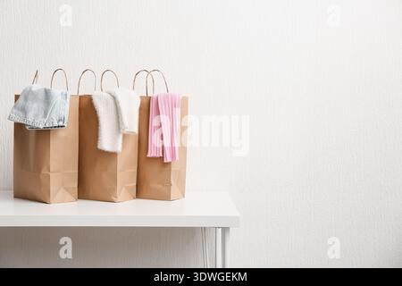 Paper shopping bags with different sweaters and jeans on table against white wall Stock Photo