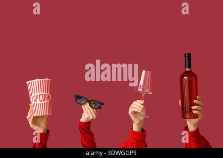 Female hands with wine glass, bucket of popcorn, 3D glasses and bottle of wine on red background. Valentine's Day celebration Stock Photo