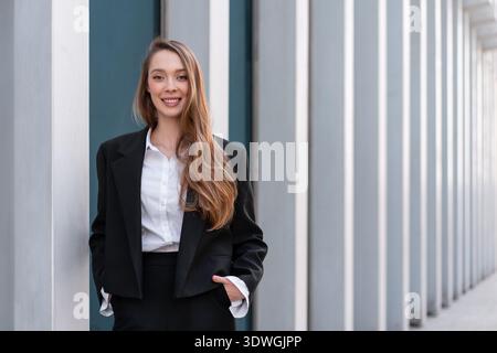 Smiling businesswoman in black suit standing against modern architectural columns. Professional confidence and corporate success concept Stock Photo