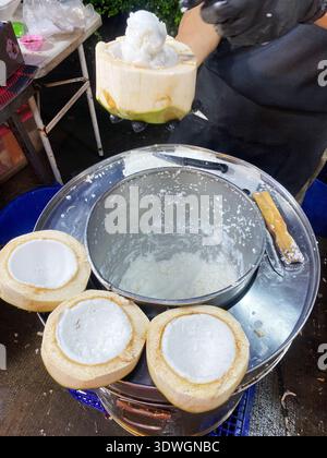 Coconut ice cream is being scooped into a coconut shell, with several empty coconut shells placed on a metal container, highlighting a street food set Stock Photo