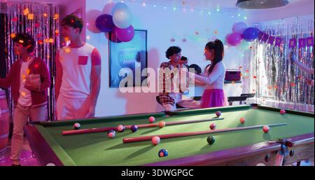 Handing woman wearing pink skirt passing trophy to seated man on pool table at birthday party Stock Photo