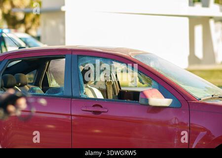 Close-up view of a red passenger car with a shattered side window parked on a city street under bright sunlight during the day. Stock Photo