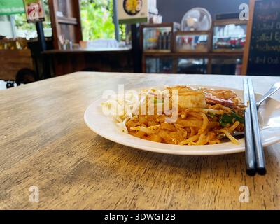 Plate of Pad Thai featuring noodles, shrimp, and bean sprouts on a wooden table, with a casual restaurant interior visible in the background Stock Photo