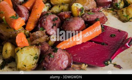 Sliced sausages and roasted vegetables are placed on a baking tray. Carrots and potatoes are mixed with herbs. The scene shows food ready to serve, hi Stock Photo