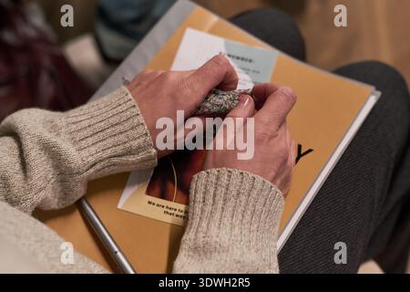 Caucasian middle aged woman holding patterned handkerchief with both hands while sitting, resting hands on stack of informational brochures, pen visible on top of papers Stock Photo