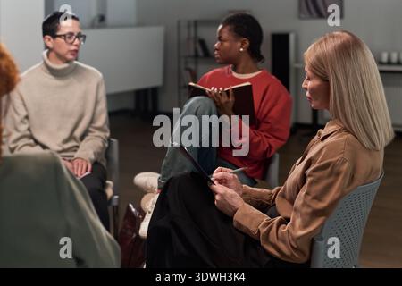 Group of diverse young adult and middle aged women sitting in circle discussing and taking notes during therapy session in modern office setting, focused on communication and collaboration Stock Photo