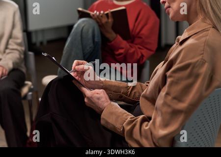 Middle aged Caucasian woman writing on clipboard during group meeting, multiethnic adults sitting in background holding notebooks, participating in discussion or therapy session Stock Photo