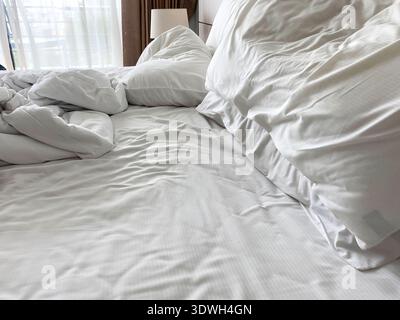 Unmade bed featuring wrinkled white linen and crumpled pillows, with soft natural light coming through sheer curtains in a comfortable bedroom Stock Photo