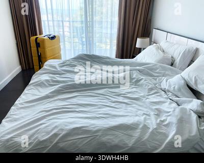 Unmade bed with wrinkled white linens and a yellow suitcase beside it in a hotel room, large window with sheer curtains providing natural light Stock Photo