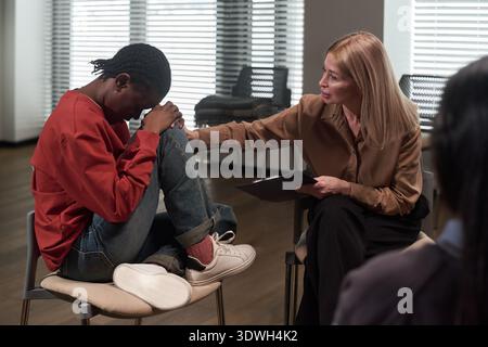 Caucasian middle aged woman comforting Black teenager sitting with knees up and head down during counseling session, therapist gently touching her arm, emotional support concept Stock Photo