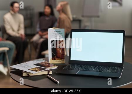 Open laptop with blank screen standing on table near brochures and notebook, background showing group of multiethnic people sitting in circle talking during therapy session Stock Photo
