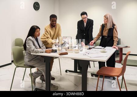 Diverse group of women collaborating around table, discussing documents and using laptops in modern office setting, engaging in teamwork and planning project Stock Photo