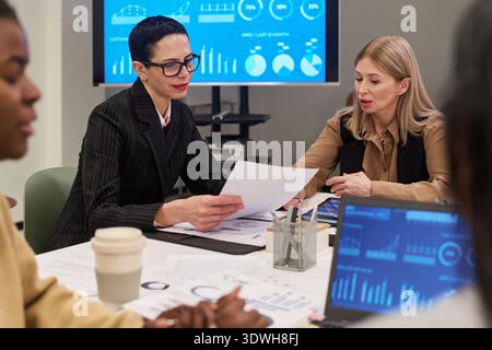 Caucasian middle aged women discussing business documents at meeting table with charts and graphs on screen, diverse colleagues collaborating nearby Stock Photo