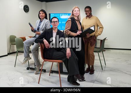 Group of four young adult and middle aged women, including Caucasian, Hispanic and Black individuals, posing confidently in modern office setting with digital screen and holding folders Stock Photo