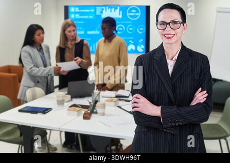 Portrait of middle aged Caucasian woman smiling with arms crossed in foreground, diverse group of women collaborating on business project with digital screen in background Stock Photo