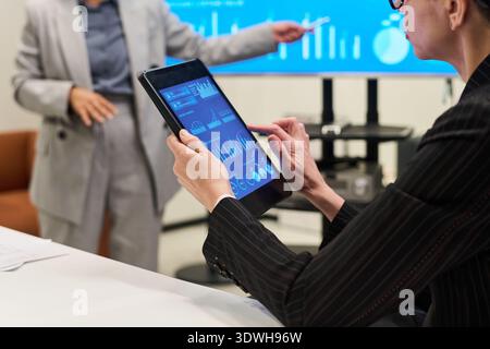 Caucasian middle aged woman using digital tablet analyzing financial data while another Hispanic woman presenting charts on screen in modern office meeting setting Stock Photo