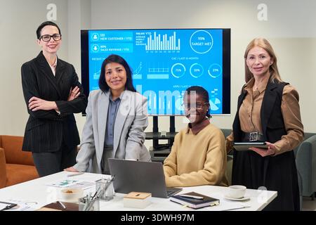Group of four businesswomen including Caucasian, Hispanic and Black women standing and sitting in modern office, smiling at camera, digital charts displayed on screen behind Stock Photo