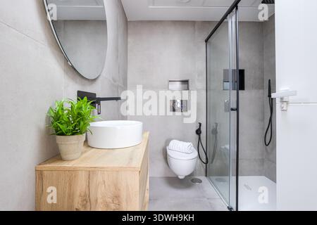 Contemporary bathroom interior featuring walk in glass shower, floating toilet, wooden vanity, round mirror, and neutral grey tile design. Stock Photo