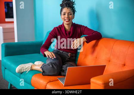 African American woman sitting on two-tone sectional sofa at home using silver laptop Stock Photo