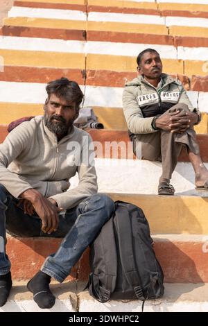 Two men sit on striped steps in Varanasi, India, with one man resting near a black backpack. Stock Photo