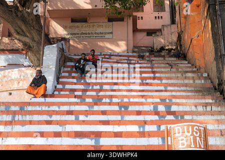 Three men sitting on orange and white painted steps beside a dustbin in Varanasi, India under a tree. Stock Photo