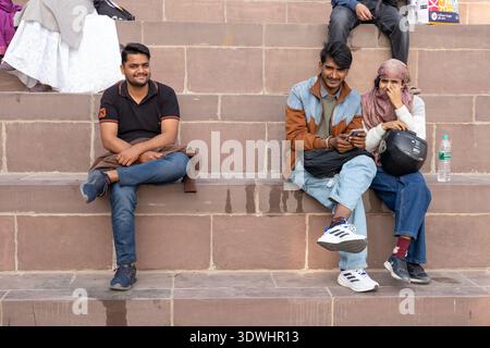 Three young adults sitting on stone steps in Varanasi, India, enjoying a casual moment outdoors. Stock Photo