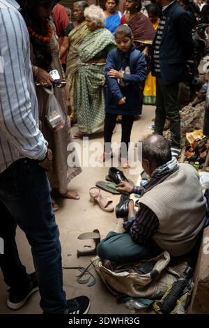 A cobbler repairs shoes on a busy street in Varanasi, India, surrounded by local people and traditional attire. Stock Photo
