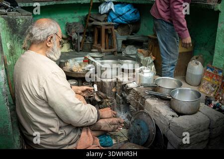 An elderly man making milk tea in a rustic kitchen setting in Varanasi, India, surrounded by cooking pots and utensils. Stock Photo