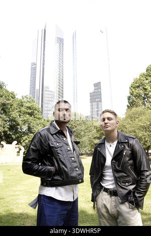 Two confident men wearing leather jackets standing in a park with tall buildings behind, outside in front of office buildings, skyscrapers, internatio Stock Photo