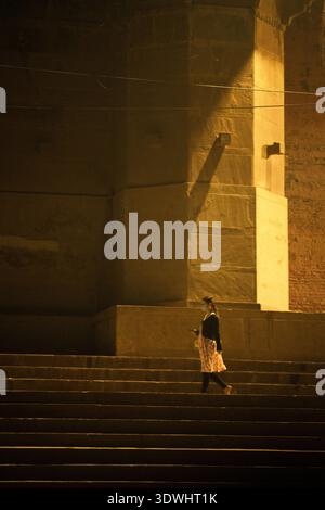 A woman wearing a face mask walks down stone steps at night in Varanasi, India, illuminated by warm light. Stock Photo