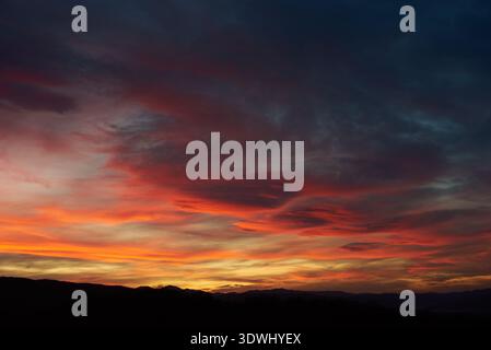 Stunning sunset paints sky with vibrant hues of red, orange, and pink, blending into deep blues and purples. Silhouetted hills create contrast against fiery horizon, adding drama to serene landscape. Stock Photo